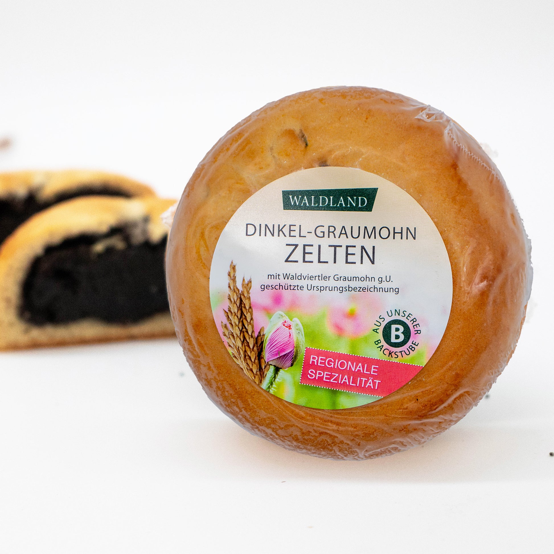 Der runde Dinkel-Graumohnzelten, in transparenter Folie verpackt liegt auf weißem Untergrund, umgeben von verstreuten Mohnsamen. Auf der Verpackung befindet sich ein Etikett mit der Aufschrift "Waldland Dinkel-Graumohnzelten – regionale Spezialität" sowie einer Abbildung von aufblühenden rosa Mohnblüten und einer Dinkelähre. Im Hintergrund sind angeschnittene Dinkel-Graumohnzelten, wo die saftige Füllung zu sehen ist.