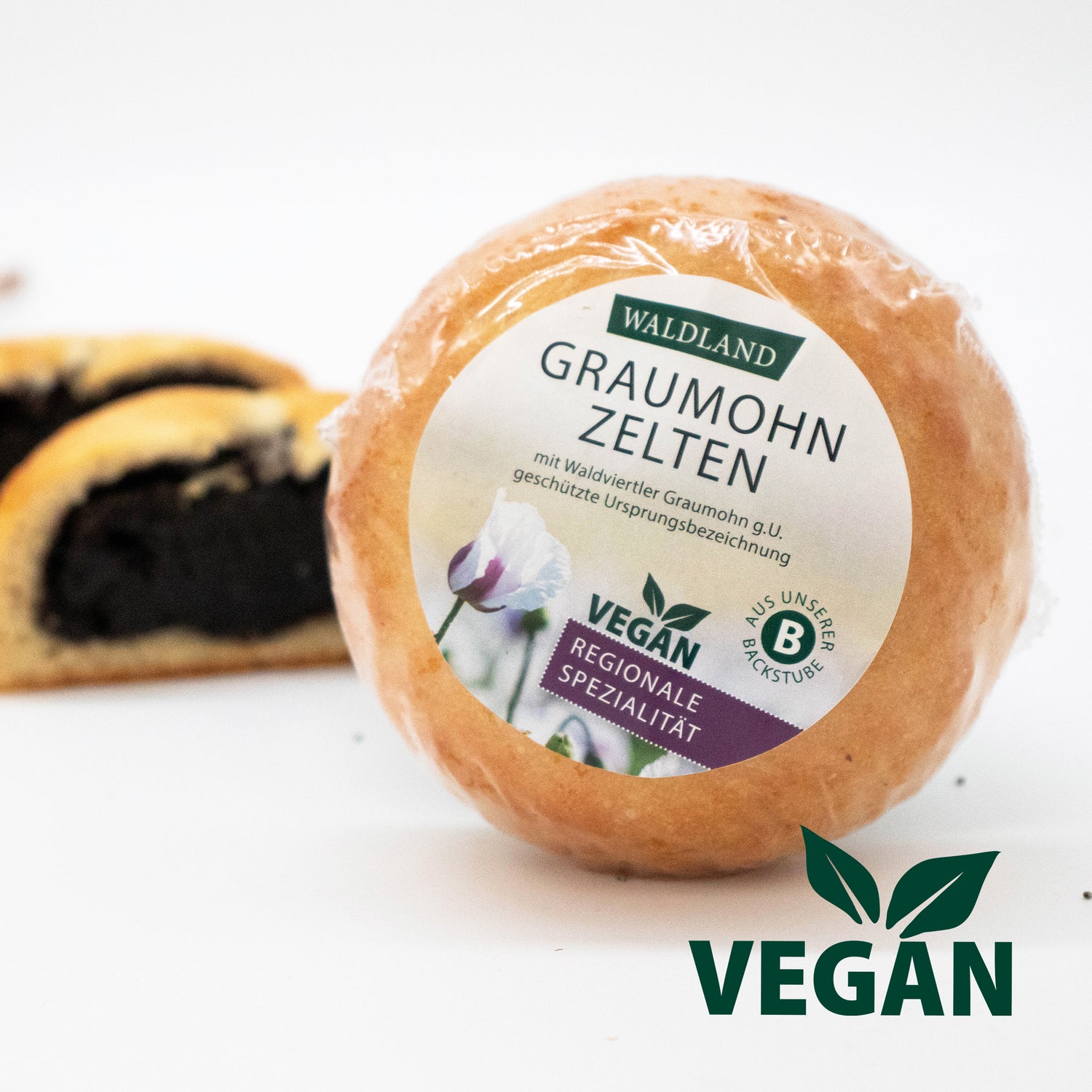 Der runde Graumohnzelten vegan, in transparenter Folie verpackt liegt auf weißem Untergrund, umgeben von verstreuten Mohnsamen. Auf der Verpackung befindet sich ein Etikett mit der Aufschrift "Waldland Graumohnzelten  vegan – regionale Spezialität" sowie einer Abbildung von einer weißen Mohnblüte. Im Hintergrund sind angeschnittene vegane Graumohnzelten, wo die saftige Füllung Graumohnfüllung zu sehen ist. Rechts unten im Bild befindet sich noch ein "VEGAN" Logo. 