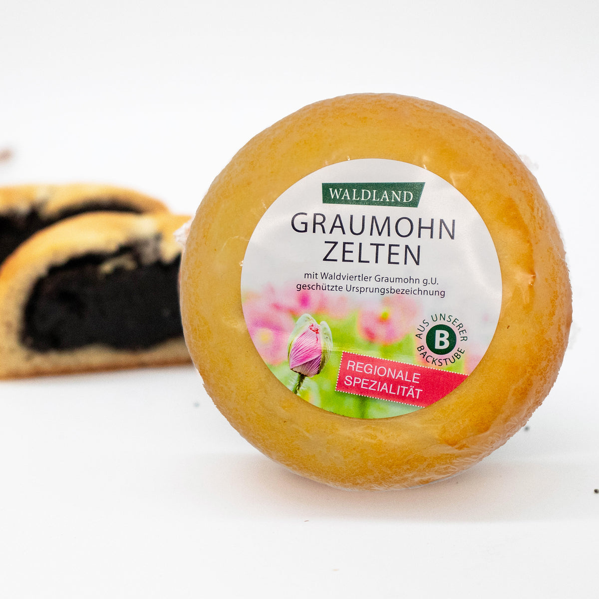 Der runde Graumohnzelten, in transparenter Folie verpackt liegt auf weißem Untergrund, umgeben von verstreuten Mohnsamen. Auf der Verpackung befindet sich ein Etikett mit der Aufschrift "Waldland Graumohnzelten – regionale Spezialität" sowie einer Abbildung von aufblühenden rosa Mohnblüten. Im Hintergrund sind angeschnittene Graumohnzelten, wo die saftige Füllung zu sehen ist.