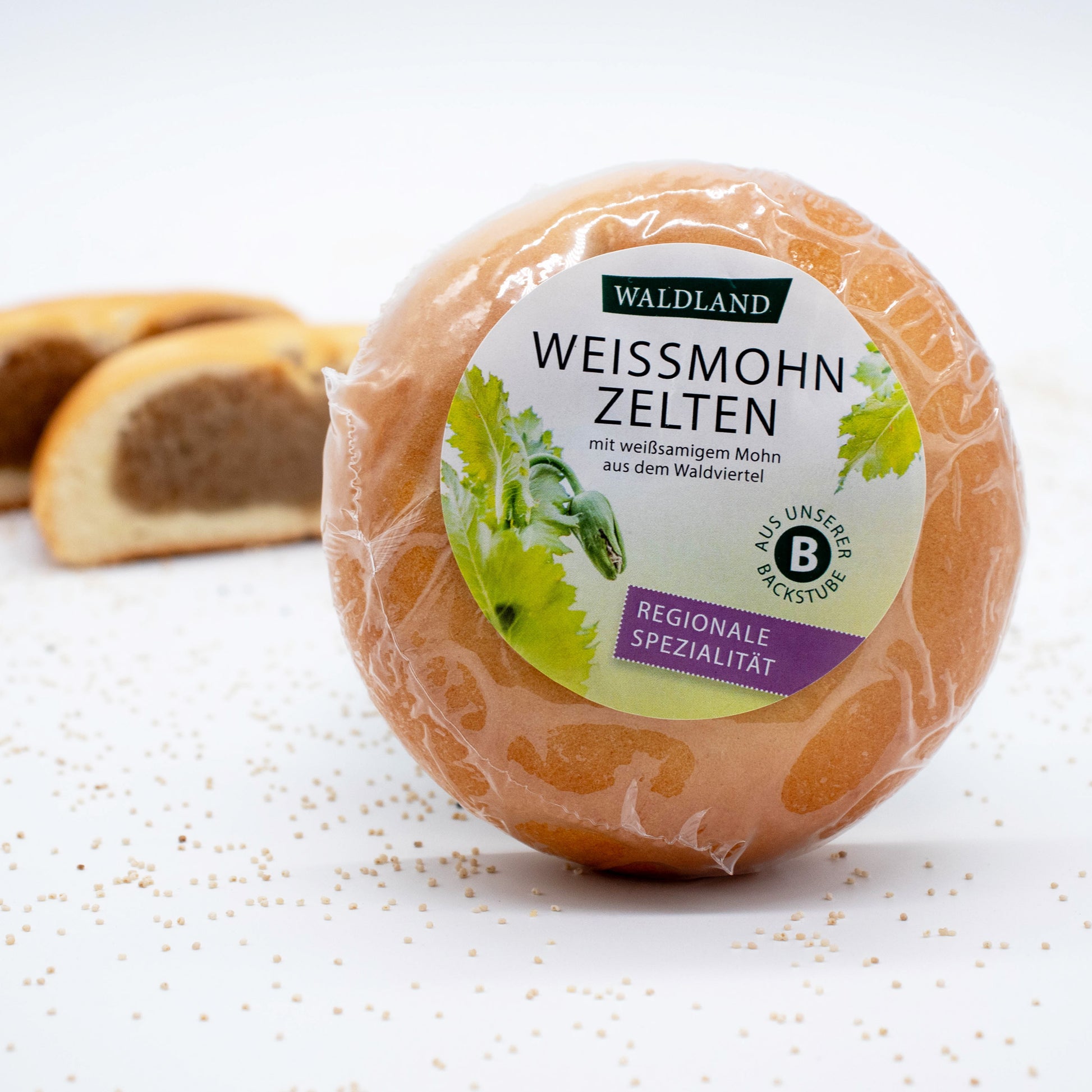 Der runde Weißmohnzelten, in transparenter Folie verpackt liegt auf weißem Untergrund, umgeben von verstreuten Mohnsamen. Auf der Verpackung befindet sich ein Etikett mit der Aufschrift "Waldland Weißmohnzelten – regionale Spezialität" sowie einer Abbildung von grünen Mohnpflanzen. Im Hintergrund sind angeschnittene Weißmohnzelten, wo die saftige Füllung zu sehen ist.